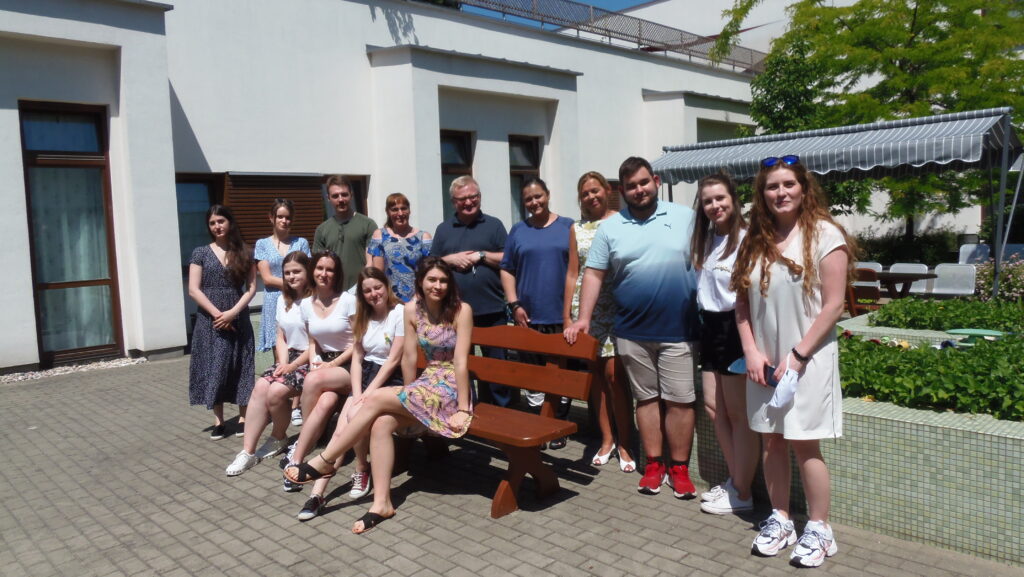 grupa studentów i pracowników Centrum Alzheimera znajduje się na słonecznym patio. Cztery studentki siedzą na ławce, pozostałe osoby stoją za nimi. W tle widać budynki oraz zieleń i parasol przeciwsłoneczny