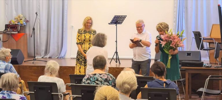 Dyrektor Centrum Alzheimera (mężczyzna)oraz zastępca dyrektora Centrum Alzheimera (kobieta) wręczają skrzypaczce dyplom z podziękowaniami za występ oraz bukiet kwiatów. Przed nimi siedzą widzowie.