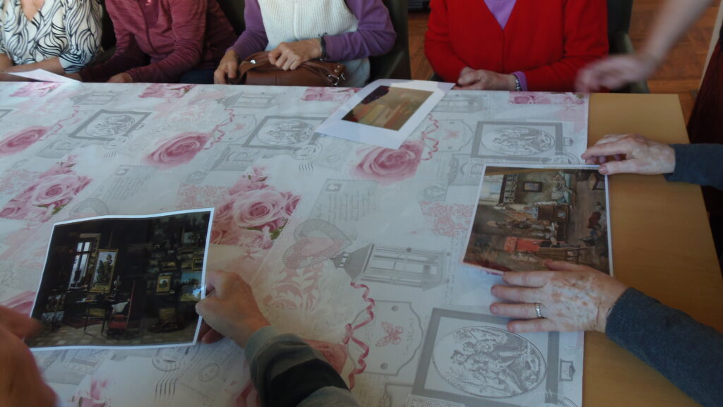 Grupa seniorów siedzi przy stole. W dłoniach trzymają reprodukcje obrazów.