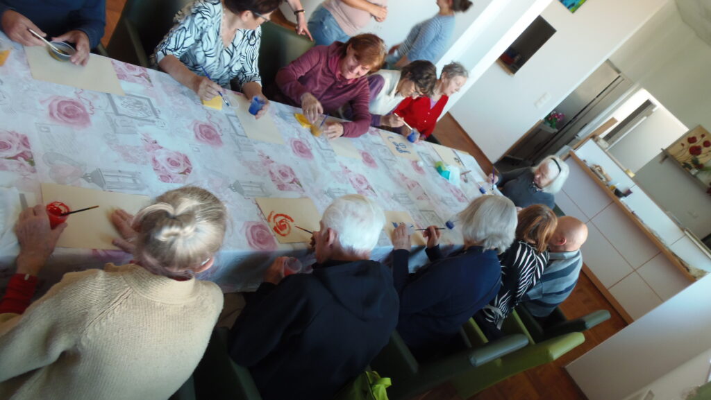 grupa seniorów siedzi przy stole pochylona nad kartonami papieru.