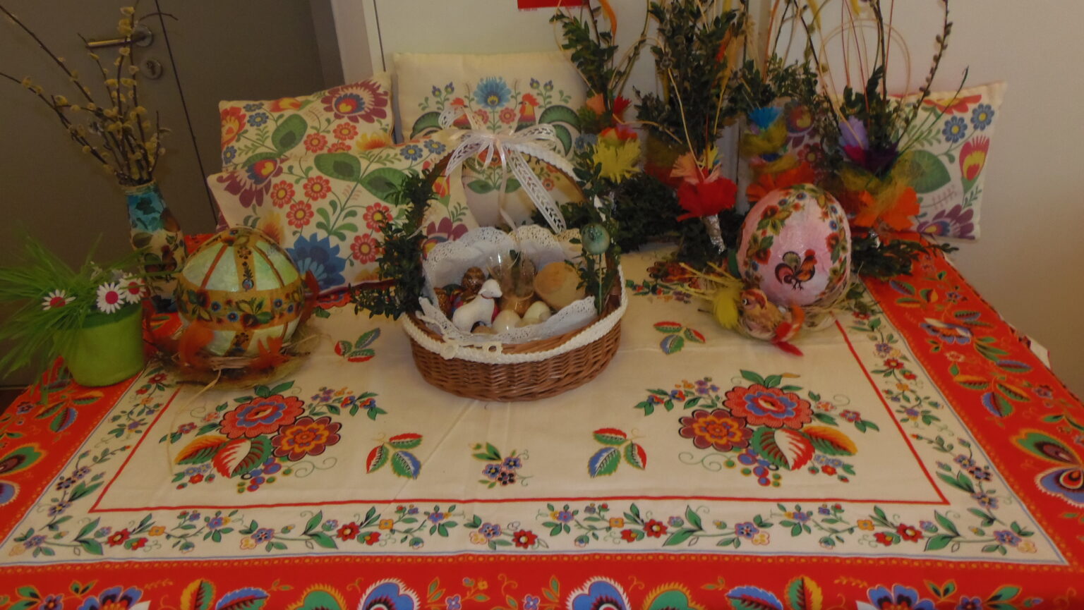 na stole przykrytym barwnym obrusem stoi koszyczek wielkanocny i ozdoby wielkanocne palmy, pisanki i kolorowe poduszki.
