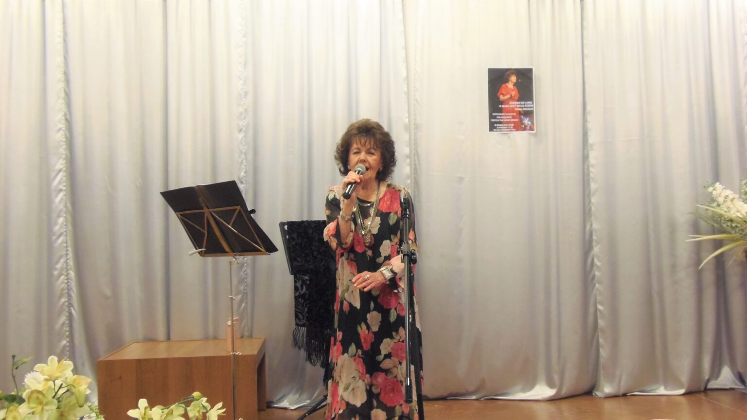 Pani Hanna Michalik podczas występu na scenie sali widowiskowej Centrum Alzheimera. Kobieta ubrana w długą suknie w kwiaty trzyma mikrofon i śpiewa.